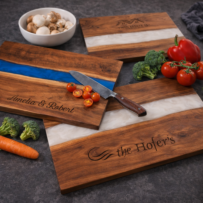 Wooden serving boards with resin river with engraved names and logos, surrounded by vegetables on a dark surface.